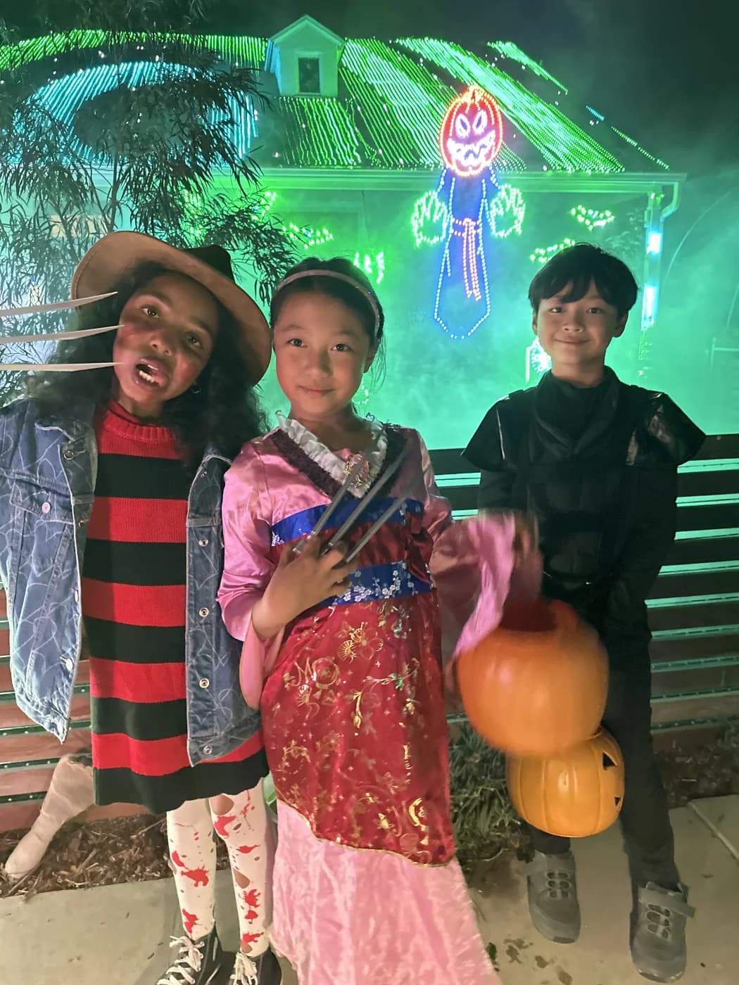 Three children in Halloween costumes smiling together in front of the Lights on Falcon display.